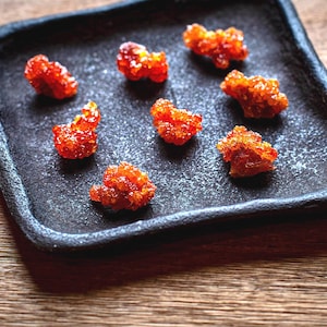 Può includere: Un piatto quadrato nero contiene diversi pezzi di cibo cristallizzato arancione brillante. Il cibo ha una forma irregolare e strutturata. Il piatto è su una superficie di legno, con il cibo come punto focale.
