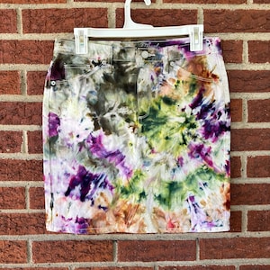 May include: A denim skirt with a vibrant tie-dye pattern in shades of purple, green, brown, and white. The skirt is hanging on a white hanger against a brick wall. The skirt has pockets and a button closure.