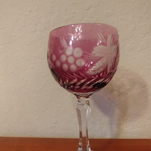 May include: A clear glass wine goblet with a deep pink hue. The goblet is etched with a grapevine design and has a long, slender stem.