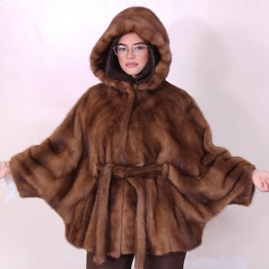 Può includere: Mantella di pelliccia marrone con cappuccio e cintura abbinata. La mantella è realizzata in morbida e lussuosa pelliccia e ha un design fluido. La modella indossa occhiali e la mantella è di un colore caldo e neutro.