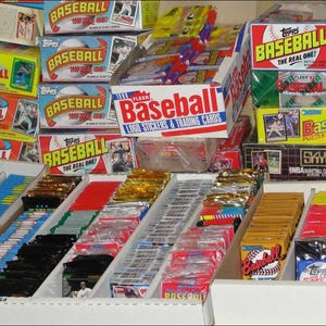 May include: A display of vintage baseball trading cards and packs. Boxes and packs feature the "Topps Baseball" and "Baseball" logos. The display includes various colors and designs, showcasing the collectible nature of the items.