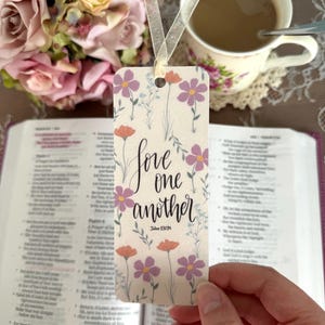 May include: A rectangular paper bookmark featuring a cream background with a floral pattern of purple and orange flowers. The text "love one another" is printed in black script in the center, with "John 13:34" below. A sheer white ribbon is attached.