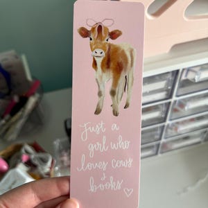 May include: Pink bookmark with a watercolor illustration of a brown and white calf with a pink bow. The text reads "Just a girl who loves cows & books".