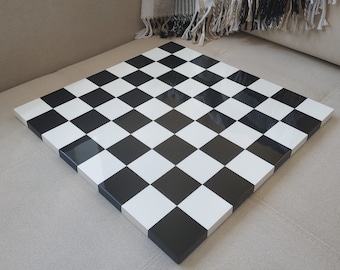 May include: A square, glossy chessboard with alternating black and white squares. The board is set on a neutral-toned surface, with a black and white patterned throw blanket in the background. A classic game board.