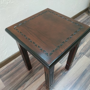 May include: A dark brown wooden end table with a square top and four legs. The top has a black border with a carved design of triangles.