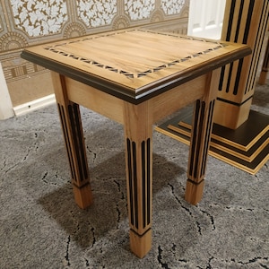 May include: A small wooden end table with a square top and four legs. The top is made of light wood with a dark wood border and a geometric pattern. The legs are also made of light wood with dark wood accents.