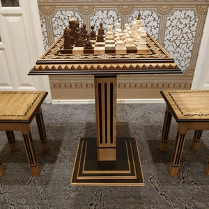 Puede incluir: Una mesa de ajedrez de madera con un tablero de ajedrez blanco y negro, tallas intrincadas y una base negra y marrón. Se incluyen dos taburetes de madera a juego con detalles negros y marrones.
