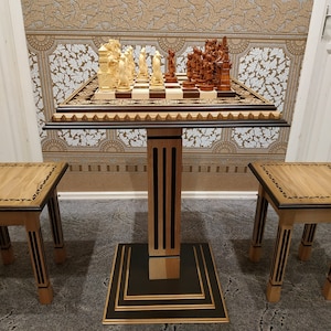 May include: A wooden chess set with intricately carved pieces on a table with two matching stools. The chessboard is black and white with a gold trim. The table and stools have a dark wood finish with gold accents.