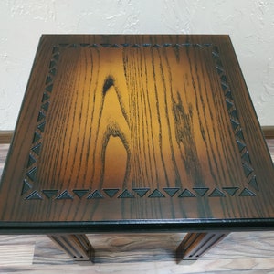 May include: A dark brown wooden end table with a black border and a carved triangular pattern. The table has a square top and four legs.