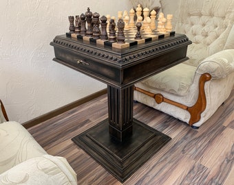 May include: A dark wood chess table with a black and white chessboard and chess pieces. The table has a decorative base and a drawer. The table is sitting on a wooden floor.