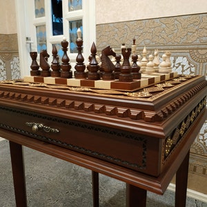 Puede incluir: Un juego de ajedrez de madera con un tablero de ajedrez a cuadros marrón y blanco, tallas intrincadas y un cajón. El juego está sobre un soporte de madera con un acabado marrón oscuro.