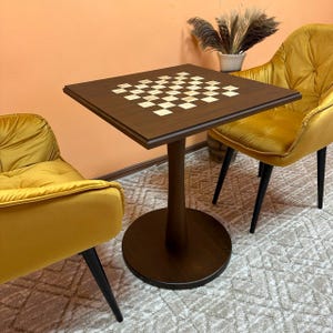 May include: A square, dark brown chess table with a checkerboard pattern on the tabletop. The table has a single, round pedestal base and is surrounded by two yellow velvet chairs with black legs. The table is approximately 24 inches square.