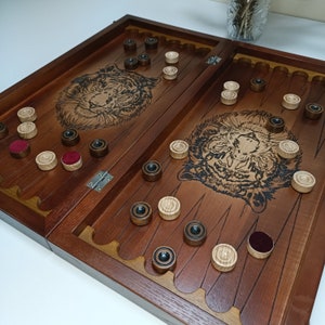Könnte beinhalten: Ein Backgammon-Brett aus dunkelbraunem Holz mit eingeätzten Tigergesichtern. Das offene Brett zeigt helle und dunkle Spielsteine, einige mit rotem Filz. Die Spielsteine sind auf dem Brett verteilt.