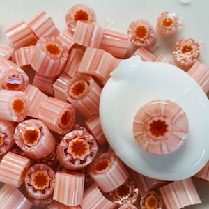 Op de afbeelding: Een verzameling roze en oranje glazen kralen met een wit bloemenontwerp. De kralen zijn op een witte ondergrond in een hoop gelegd.