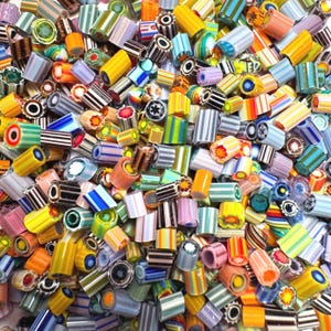 May include: A close-up view of a pile of colorful glass beads. The beads are cylindrical, with various patterns and colors, including stripes, swirls, and floral designs. The colors include yellow, orange, blue, green, and white.