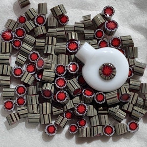 May include: A white ceramic bottle stopper with a red and silver design. The stopper is surrounded by a pile of small, red and silver striped glass beads.