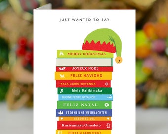 May include: A Christmas card featuring a stack of books with festive greetings in various languages, topped with an elf hat. The words "JUST WANTED TO SAY" are printed at the top of the card.