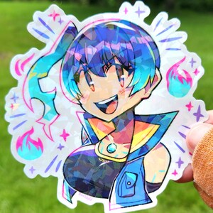 May include: A holographic sticker featuring a smiling anime-style character with blue and purple hair. The character wears a blue and yellow outfit with a gem. The sticker has a starburst design with pink and blue flames and stars.