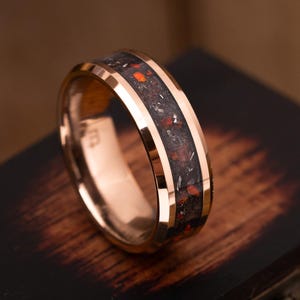 May include: A rose gold-toned ring featuring a beveled edge and a central inlay of dark, textured material speckled with vibrant orange and metallic silver flecks. The band has a polished finish and a smooth interior surface with a small engraved mark.