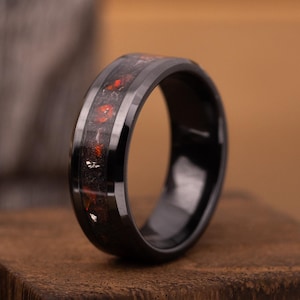 May include: A black ring with a band inlaid with red and silver flecks. The ring is set on a small, dark wooden block. The ring's band has a glossy finish, and the inlay is visible across the top of the ring.