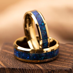 May include: Two gold rings with a blue and green opal inlay. The rings are displayed on a wooden block, highlighting their polished finish and detailed design. The opal inlay offers a striking contrast against the gold.