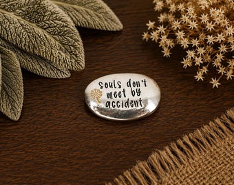 May include: A smooth, oval-shaped silver-toned metal pocket stone featuring the engraved black text "souls don't meet by accident" and a small gold-colored dandelion graphic. The object has a polished, reflective surface and rounded edges.