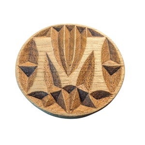 May include: A round, wooden coaster with a geometric design. The coaster features a central, vertical design with leaf-like shapes and triangular accents. The wood has a natural, light brown color with darker brown carved details.