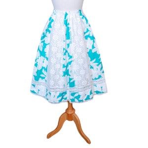 Puede incluir: Una falda floral blanca y turquesa con ribete de encaje. La falda está en un maniquí de madera.