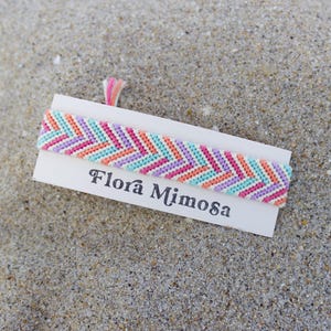 May include: A woven friendship bracelet featuring a chevron pattern in shades of light purple, turquoise, and coral pink. The bracelet is attached to a white rectangular display card with the text Flora Mimosa printed in black lettering.