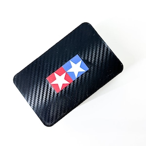 Peut inclure: Étui rectangulaire noir avec une texture en fibre de carbone. Un petit emblème avec deux étoiles blanches sur des carrés rouges et bleus est centré sur l'étui. L'étui est probablement destiné à ranger de petits objets.