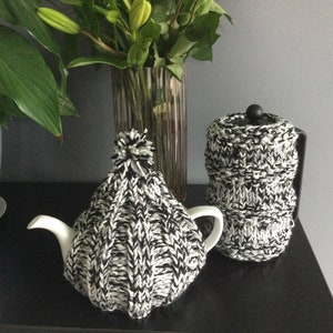 May include: A white teapot and a black French press, both with black and white knitted cosy covers. The teapot has a pom-pom on top.