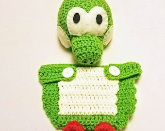 May include: A crocheted baby outfit in green and white with a matching hat and red booties. The hat features a green dinosaur head with white eyes and a red crest.