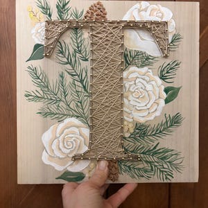 May include: Wooden wall art featuring a large letter "T" crafted with gold string, surrounded by painted white roses, green pine needles, and a small pinecone. The background is a light wood grain.