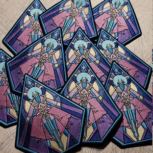 May include: A stack of colorful embroidered patches featuring a stylized figure in a stained glass design. The figure is wearing a pink and blue robe and holding a sword. The patches are made of fabric and have a black border.