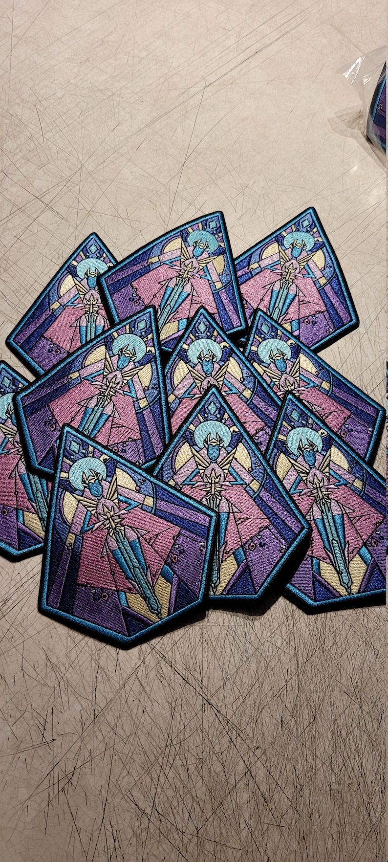 May include: A stack of colorful embroidered patches featuring a stylized figure in a stained glass design. The figure is wearing a pink and blue robe and holding a sword. The patches are made of fabric and have a black border.
