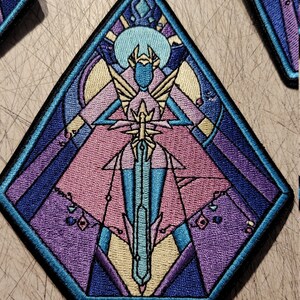 May include: A blue, purple, and pink embroidered patch with a geometric design. The patch features a figure holding a sword, surrounded by a crescent moon and stars.