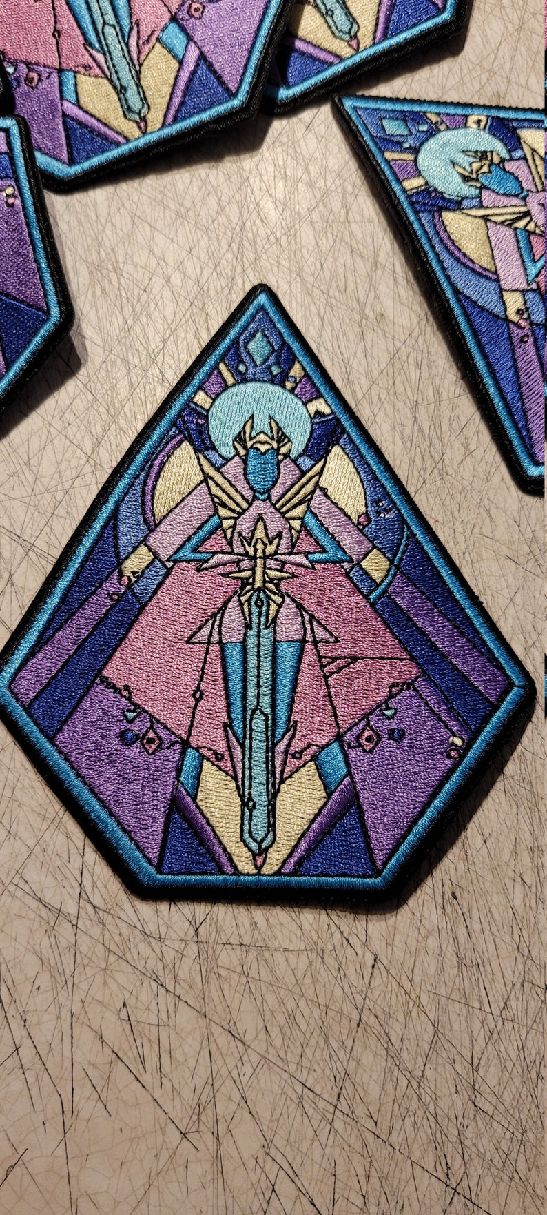May include: A blue, purple, and pink embroidered patch with a geometric design. The patch features a figure holding a sword, surrounded by a crescent moon and stars.