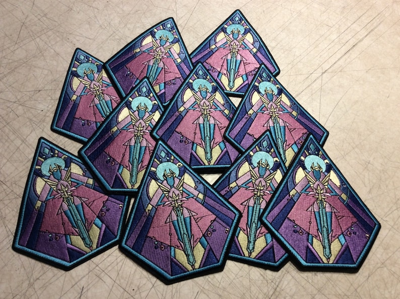May include: A collection of ten embroidered patches featuring a stylized figure in a pink and blue gown with wings. The figure is surrounded by a geometric pattern in shades of purple, blue, and yellow.
