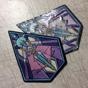 May include: Two embroidered patches with a geometric design in shades of purple, blue, and yellow. The patches are shaped like a diamond and a hexagon.