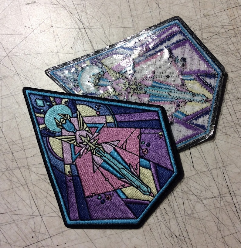 May include: Two embroidered patches with a geometric design in shades of purple, blue, and yellow. The patches are shaped like a diamond and a hexagon.