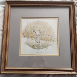 May include: Framed artwork featuring a pen and ink drawing of a lamb in a field. The lamb is white with a light brown background of trees and grass. The artwork is signed and framed in a brown wooden frame with a gray mat.