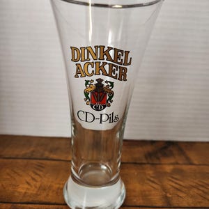 May include: A clear, tall beer glass with a gold rim. The glass features the words "DINKEL ACKER" in gold lettering, along with a crest and the text "CD-Pils". The glass is empty and sits on a wooden surface.