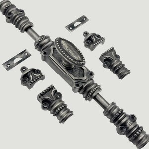 Op de afbeelding: Een zilverkleurige metalen raamgrendelset met een centrale ovale knop, een lange verticale stang met decoratieve geribbelde secties, twee rechthoekige sluitplaten met boogvormige uitsparingen en vier bijpassende montagebeugels met schroefgaten.