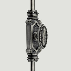 Op de afbeelding: Een donker metalen raamgrendel met een ovaalvormige knop met een getextureerd, geput oppervlak. Het mechanisme is gemonteerd op een verticale stang en bevat een rechthoekige basisplaat met vier zichtbare schroeven en decoratieve geribbelde uiteinden.