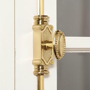 May include: Close-up of a gold window latch. The latch features an oval handle with a textured surface and a rectangular base with decorative detailing. The latch is mounted on a white surface.
