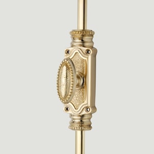 Op de afbeelding: Een raamgrendel van gepolijst messing met een ovale knop met een textuur. De centrale plaat heeft een decoratieve rand en een gestippelde afwerking, met montagegaten aan de boven- en onderkant. Het beslag is bevestigd aan een verticale stang.