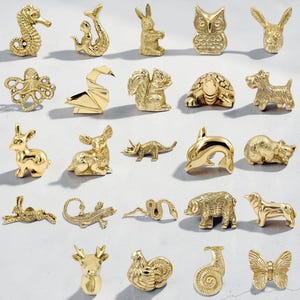 Könnte beinhalten: Eine Sammlung kleiner, goldfarbener Tierfiguren, darunter ein Seepferdchen, eine Meerjungfrau, ein Kaninchen, eine Eule und ein Hund. Weitere Tiere sind ein Oktopus, ein Schwan, ein Eichhörnchen, eine Schildkröte und ein Delfin. Die Figuren sind auf einer hellgrauen Oberfläche ausgestellt.