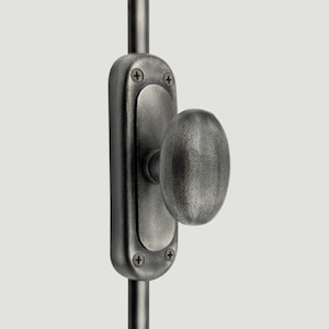 Op de afbeelding: Een donkergrijze metalen raamgrendel met een ovale knop en een rechthoekige montageplaat. Het beslag is bevestigd met twee zichtbare schroeven aan de boven- en onderkant, gemonteerd op een verticale metalen stang met een getextureerde afwerking.