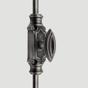 Op de afbeelding: Een donkergrijze metalen raamgrendel met een ovaalvormige handgreep en een rechthoekige basisplaat met een textuur in het midden. Het beslag is bevestigd met vier zichtbare schroeven en gemonteerd op een verticale metalen stang.
