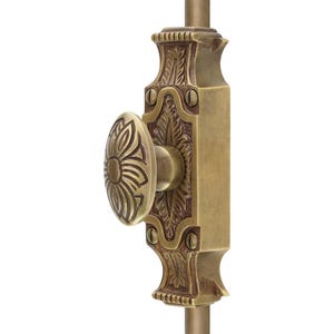 Op de afbeelding: Een messingkleurige deursluiting met een ovale handgreep met een bloemmotief. De rechthoekige achterplaat en handgreep zijn versierd met bladeren. De hardware is op een verticale staaf gemonteerd.